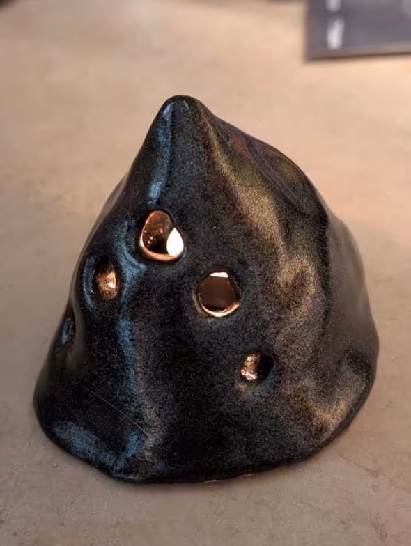  Handmade ceramic candle holder with a dark iridescent glaze and cone-like shape, featuring several cut-out holes of varying sizes 