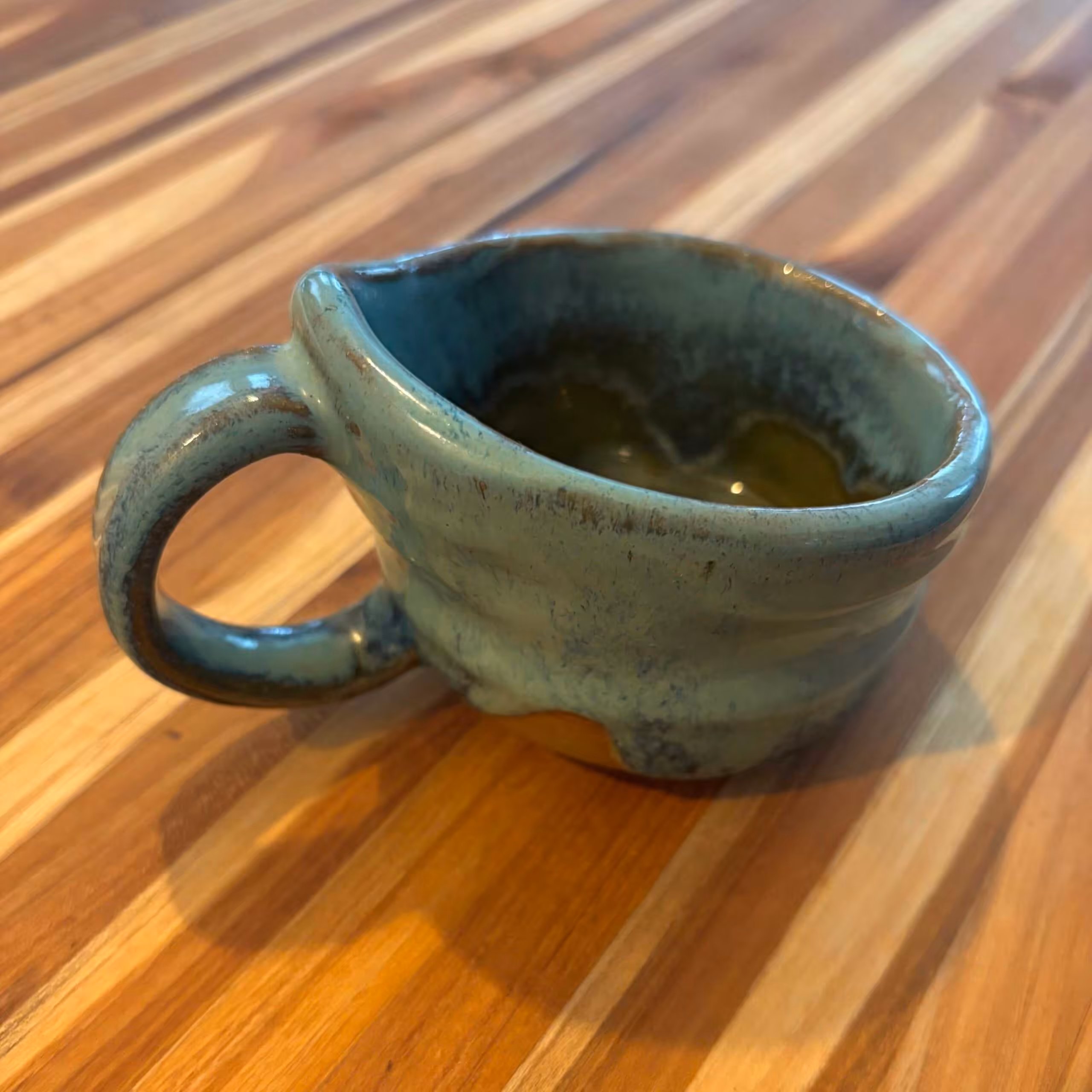  Handmade ceramic mug with a blue-green glaze and organic wavy form, sitting on a striped wood surface. The glaze shows natural variation with darker tones around the rim and lighter areas on the textured ridges. 
